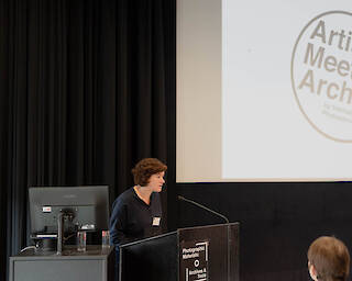 Inga Schneider steht an einem Sprechpult. Hinter ihr an einer Leinwand sieht man das Logo von Artist Meets Archive.