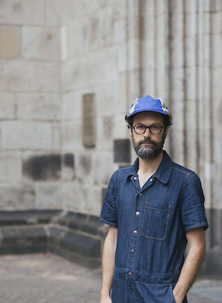 Andres Galeano in einem Jeans-Overall, mit einer Cappy und Brille. eine Hand in der Hosentasche.