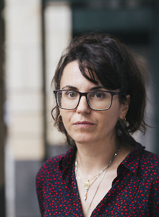 Portrait von Elena Efeoglou. Sie hat lange dunkle Haare und trägt eine Brille und eine dunkle Bluse mit roten Punkten.