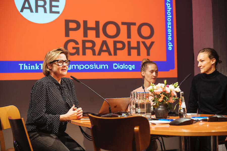 An einem runden Tisch sitzen 3 Personen mit Mikrophon in einer Diskussion. Hinter ihnen ist eine Leinwand, auf der steht We Do/Are Photography.