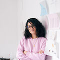 Portrait of Btihal Remli. She is wearing glasses and a pink sweater and is leaning against the wall of her studio.