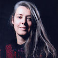 Portrait of Marta Bogdanska. She has long hair streaked with gray and is wearing a red and purple patterned sweater.