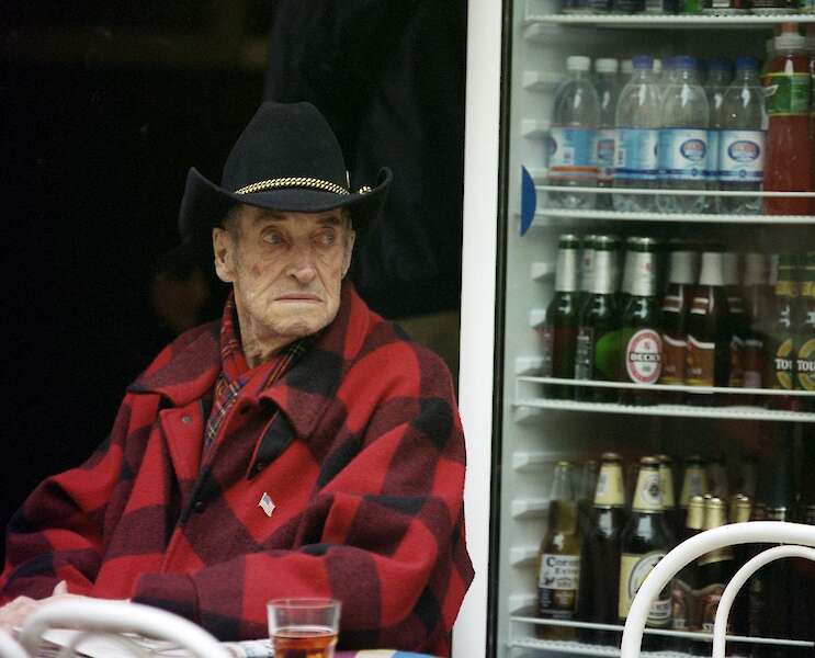 Ein älterer Herr mit einem schwarzen Cowboy Hut sitzt draußen neben einem Kühlschrank. Durch die Scheibe des Kühlschranks kann man verschiedene Flaschengetränke sehen. Vor ihm auf einem Tisch steht ein volles Glas mit einem dunklen Getränk.