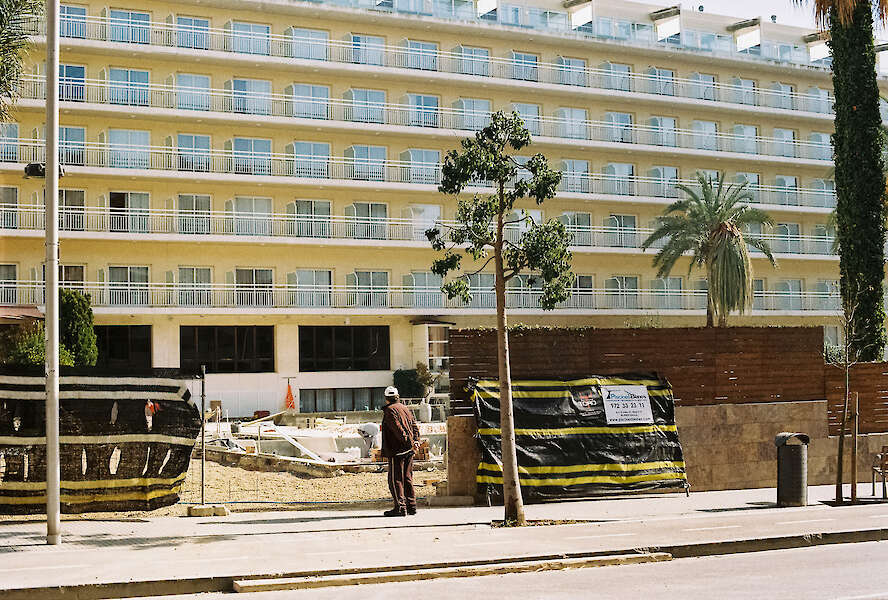 [Eine Person bleibt vor einem sehr großen Hotel in dem Urlaubsort Lloret de Mar, Mallorca, stehen. Die Hotelfassade füllt das gesamte Bild aus und zeigt sechs Stockwerke. Sichtbar sind zwanzig aneinander gereihte Fenster und Balkone. Das Hotel ist geschlossen. Ein Bauzaun grenzt das Grundstück ab. An dem Zaun hängt ein Schild mit der Telefonnummer einer Firma.]