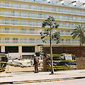 [Eine Person bleibt vor einem sehr großen Hotel in dem Urlaubsort Lloret de Mar, Mallorca, stehen. Die Hotelfassade füllt das gesamte Bild aus und zeigt sechs Stockwerke. Sichtbar sind zwanzig aneinander gereihte Fenster und Balkone. Das Hotel ist geschlossen. Ein Bauzaun grenzt das Grundstück ab. An dem Zaun hängt ein Schild mit der Telefonnummer einer Firma.]