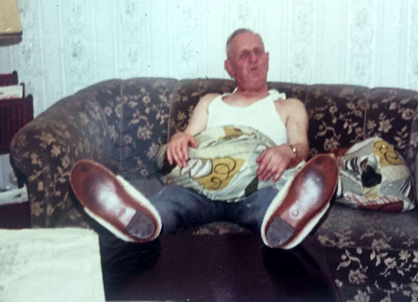 Ein älterer Mann in Unterhemd und Hausschuhen sitzt entspannt auf einem gemusterten Sofa, die Beine ausgestreckt, mit einem Kissen auf dem Schoß. Wohnzimmerambiente der 1970er- oder 1980er-Jahre.